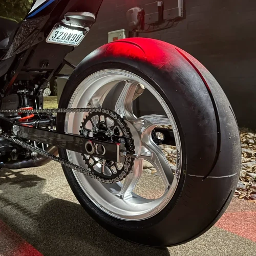 Vortex Motorcycle Parts Vortex V4 Lightweight Rear Sprocket, close-up view of the motorcycle's rear wheel and sprocket with chain, mounted on a sleek black motorcycle with a reflective tire, outdoor nighttime setting.