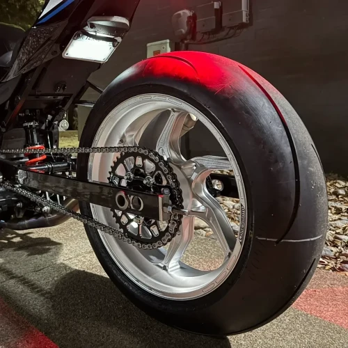 Vortex Motorcycle Parts Vortex V4 Lightweight Rear Sprocket, close-up view of the motorcycle's rear wheel and sprocket with chain, mounted on a sleek black motorcycle with a reflective tire, outdoor nighttime setting.