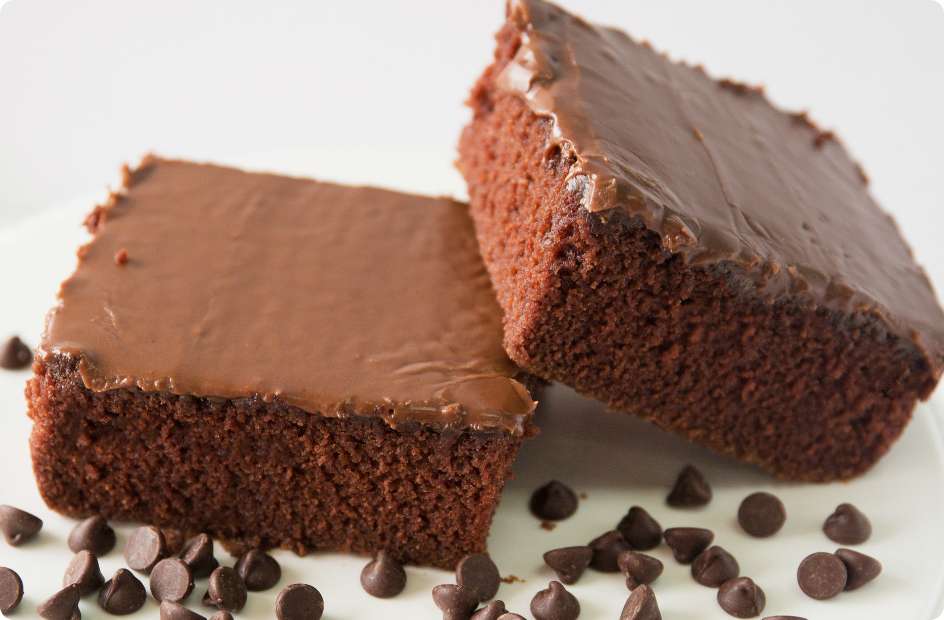 Two slices of Chocolate Texas Sheet Cake stacked on a plate with chocolate chips scattered around the base of the plate