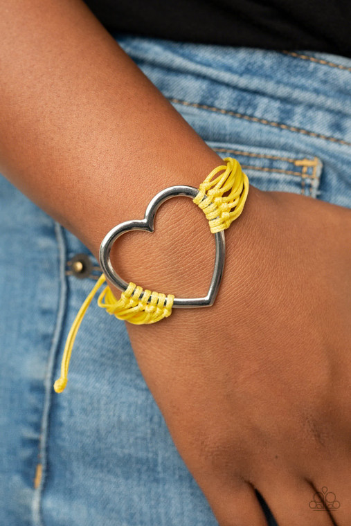 Layers of yellow cords delicately knot around an oversized silver heart charm, creating a whimsical centerpiece around the wrist. Features an adjustable sliding knot closure.

Sold as one individual bracelet.