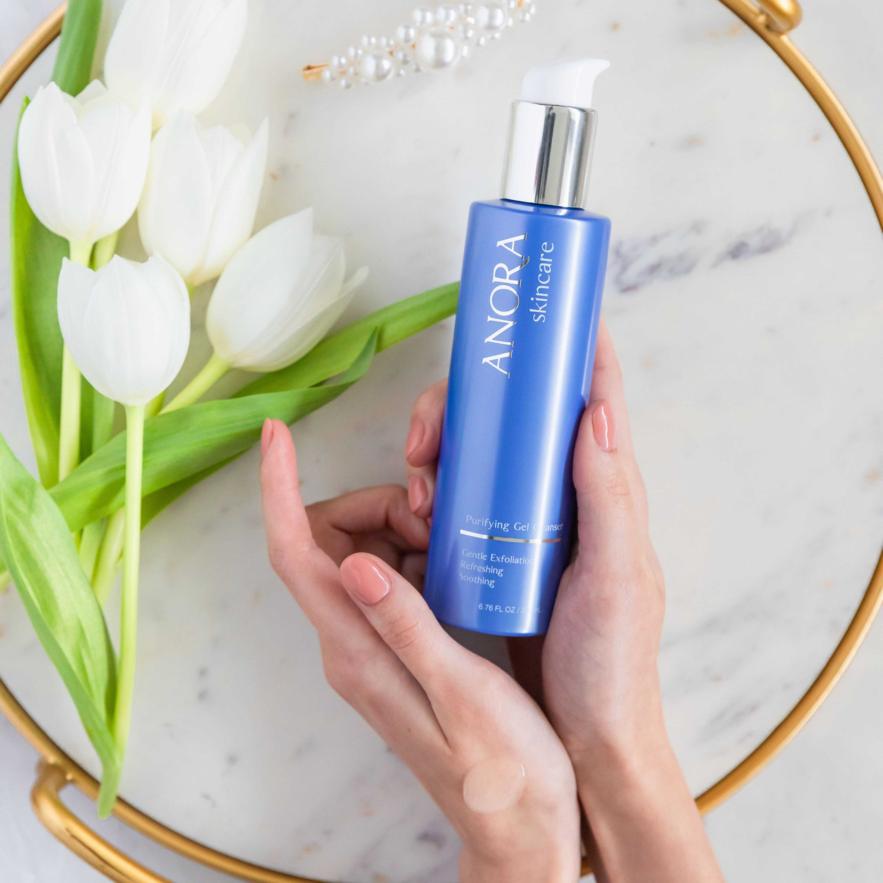 Hands holding Anora Gel Cleanser over a marble tray with white tulips
