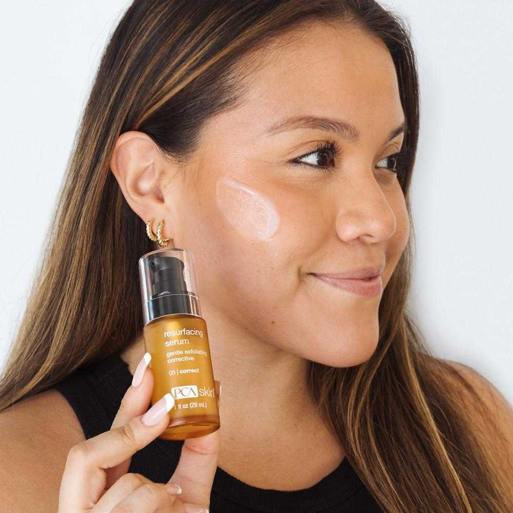 Smiling woman holding Resurfacing Serum with product on her cheek