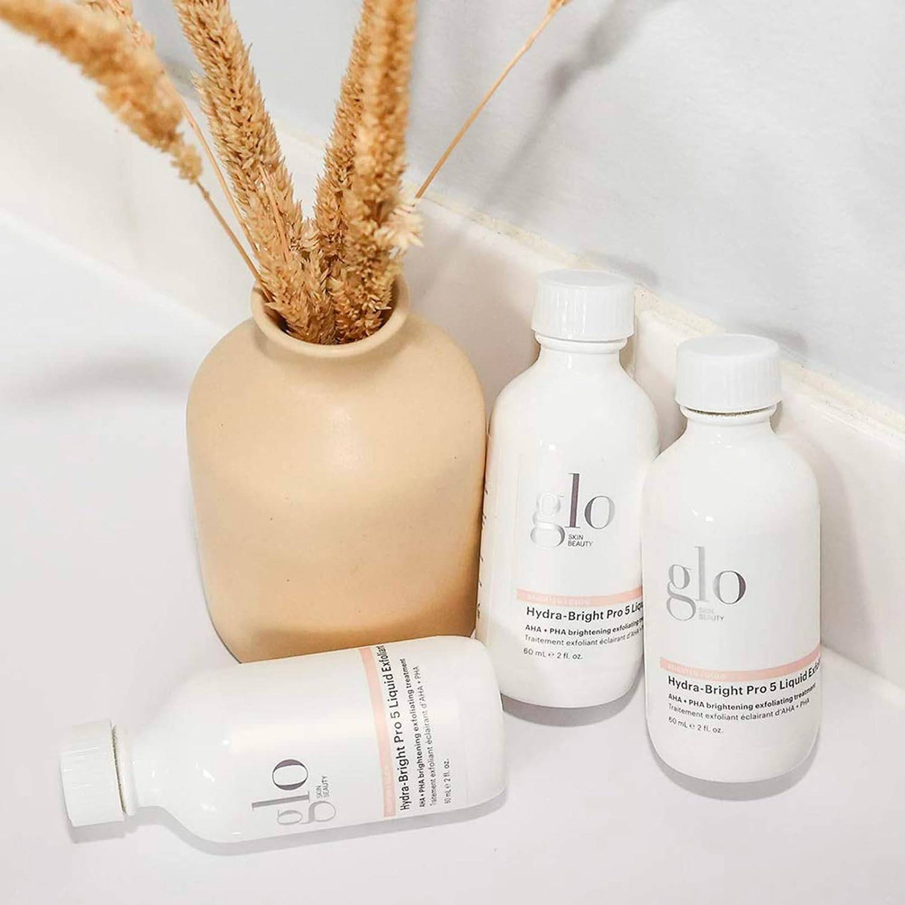 Three glo Hydra-Bright exfoliant bottles beside a vase of dried grasses