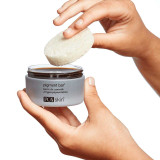 Hands lifting a beige Pigment Bar cleansing bar from its open jar