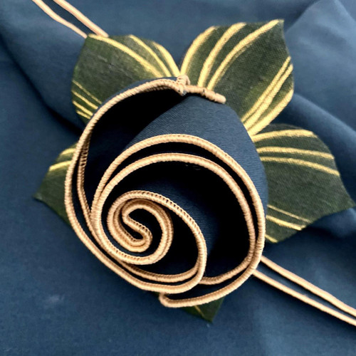 Pine with Gold Trim Napkin, intricately rolled navy blue napkin with gold trim detail, complimented by green leaves background, designed by Carole Shiber.