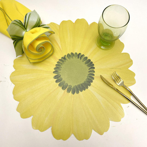 Gerber Daisy Placemat, Yellow Carole Shiber Designs, yellow flower-shaped placemat with a green center, placed on a white surface with a green glass, a yellow cloth napkin decorated with a green ribbon, and gold-colored cutlery.