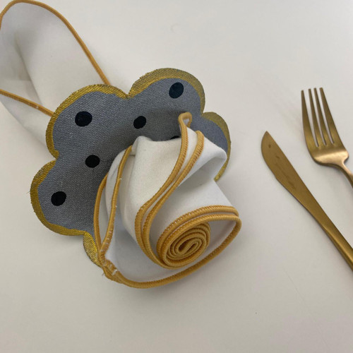 Polka-Dotl Ring-to-Coaster, Black Dots on Grey Carole Shiber Designs, a stylish grey ring-to-coaster with black polka dots and gold edges, displayed with a rolled white napkin with gold trim, alongside gold-colored cutlery on a white table.