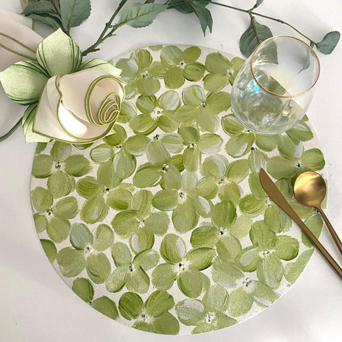 Green Petals on Round Carole Shiber Designs, a decorative round table centerpiece with painted green flower pattern, accompanied by a gold spoon and a cutlery knife, with a glass and a ribbon bow on a white surface. Green Petals on Round Carole Shiber Designs, a decorative round table centerpiece with painted green flower pattern, accompanied by a gold spoon and a cutlery knife, with a glass and a ribbon bow on a white surface.