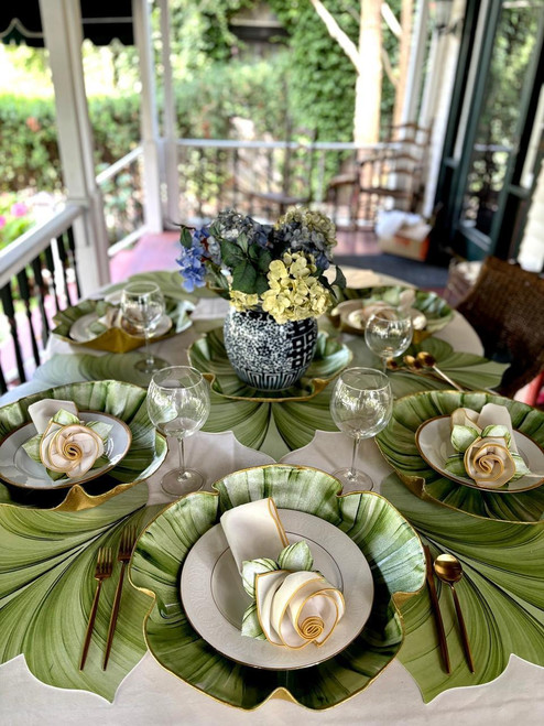Signature SculpTable Linen Botanic Tablescape by Carole Shiber Designs, a beautifully arranged table setting featuring large green leaf placemats, elegant white and gold napkins with rose-shaped details, and a central floral arrangement with white, yellow, and blue flowers in a black and white patterned vase, on a sunroom patio with lush greenery outside.