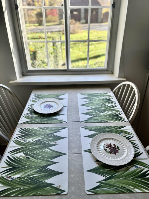 Winter Mountain Table by Carole Shiber Designs, showcasing a table with tropical leaf-themed placemats and decorative plates near a window with a garden view.