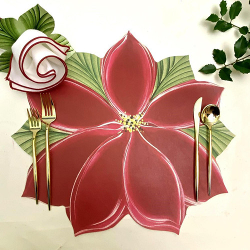 Add a Poinsettia Placemat Carole Shiber Designs, festive table setting with a large poinsettia flower decorated placemat, gold cutlery, a fabric flower napkin, and greenery in the background.