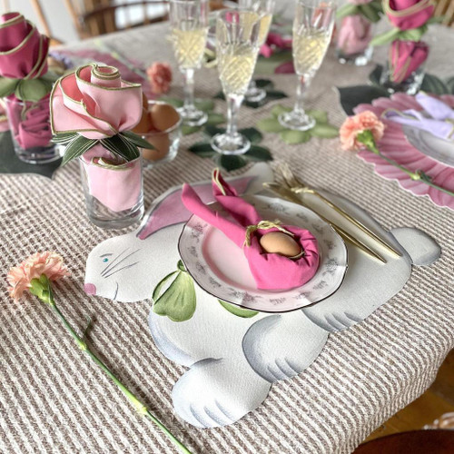 Add a Easter Bunny Placemat Carole Shiber Designs, a decorative placemat featuring a bunny illustration with floral and pastel accents, set on a tablescape with pink napkins folded into bunny shapes.