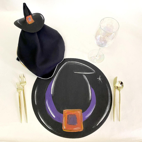 Add a Witch's Hat Placemat Carole Shiber Designs, Halloween-themed black placemat with purple and orange witch's hat design, black witch's hat with orange buckle accessory on a dining table.