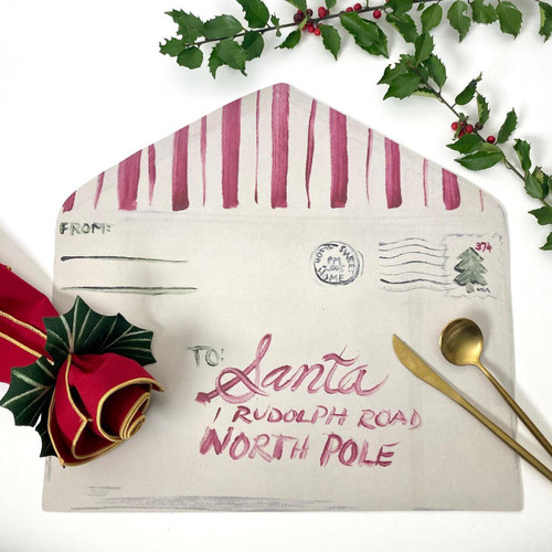 Add a Letter To Santa Placemat Carole Shiber Designs, festive holiday-themed placemat with a painted letter to Santa, surrounded by Christmas decorations including holly leaves, red berries, a gold spoon, a gold knife, and a red gift with a holly leaf embellishment.