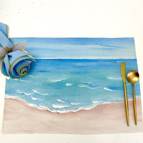 A Day at the Beach Placemat Carole Shiber Designs, watercolor painted beach scene with waves and sand, accompanied by gold-toned spoon and knife, and a fabric flower decoration.
