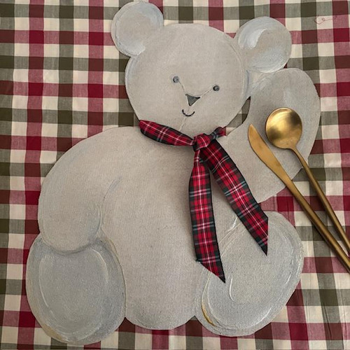 Teddy Bear Placemat with Plaid Ribbon, beige plush teddy bear-shaped placemat with red and black plaid ribbon around its neck, placed on a red and white checkered tablecloth with gold spoon and fork.