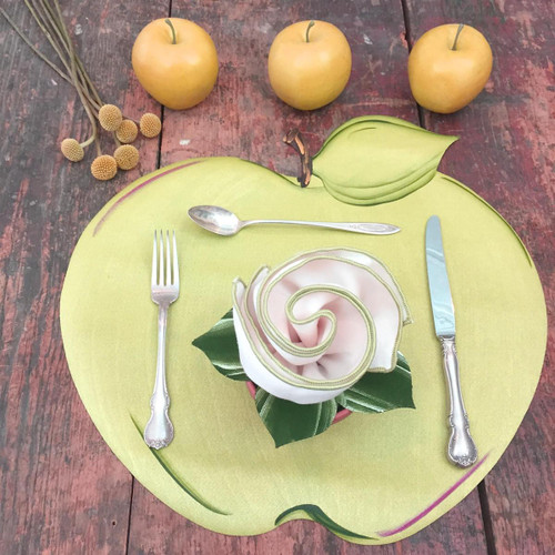 Green Apple Placemat Carole Shiber Designs, round green placemat with apple leaf design, set on rustic wooden table with colorful decor including apples and a fabric flower centerpiece.