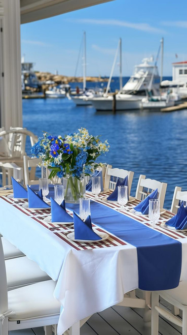 Carole Shiber Designs, custom tablecloths set at a seaside dining table with blue accents, floral centerpiece, and marina view in the background.