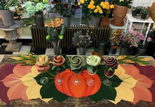 Earthtone Runner Assorted 6 Carole Shiber Designs, floral napkin arrangement with roses and greenery on a table, surrounded by colorful flowers and potted plants in a garden or flower shop setting.