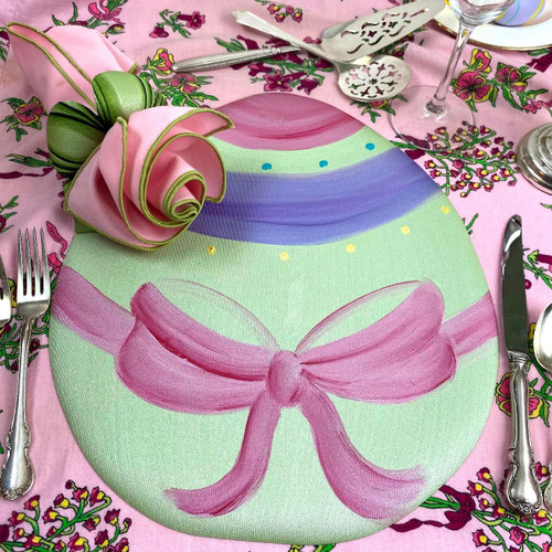 Easter Egg Placemat Carole Shiber Designs, pink with a painted pink bow and pastel rainbow stripes on a pink floral tablecloth with dinnerware and utensils.
