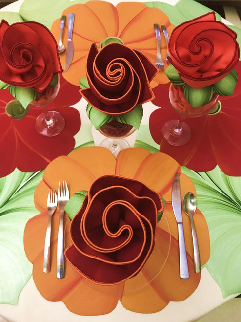 Popping Orange Poppy, LIMITED EDITION Carole Shiber Designs, elegant table setting with napkins folded into roses in orange and red, set on a floral-themed tablecloth with large orange and green flower patterns.