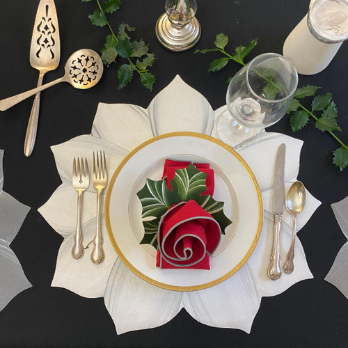 White/Silver Poinsettia Place-Setting Carole Shiber Designs, elegant holiday table setting with a poinsettia-themed napkin on a white plate with gold trim, surrounded by silverware, glassware, and festive greenery.