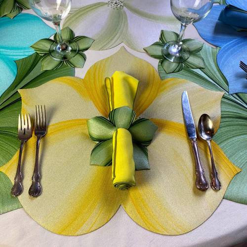 Lily Placemat, Yellow - Carole Shiber Designs; image shows a vibrant yellow lily-shaped placemat on a dining table, decorated with green and yellow napkins, glassware, and silver cutlery.