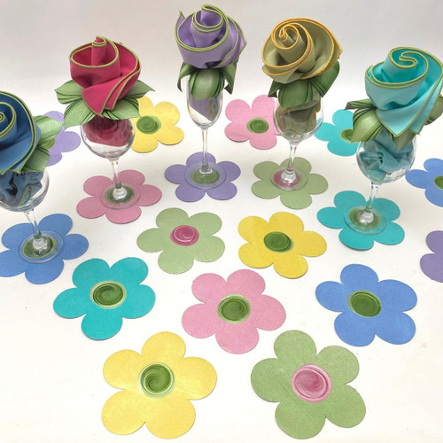 One Dozen Flower Power Coasters, colorful paper flower coasters arranged on a white surface with wine glasses decorated with fabric roses by Carole Shiber Designs.