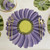 Gerbera Daisy Placemat - Lilac, close-up of a purple daisy-themed placemat with a yellow center, set on a table with gold utensils and green accents.