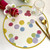 Happy Dots! Carole Shiber Designs, colorful polka dot placemat with a pink flower, gold spoon and knife, and a clear glass on a white table.