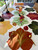 Autumn Leaves, Set for 12 by Carole Shiber Designs - colorful fall-themed tablecloth with large leaf patterns in red, green, yellow, and brown, decorated with a vase of rolled paper roses as centerpiece, gold cutlery, and black and gold candlesticks for an elegant fall dining setup. Autumn Leaves, Set for 12 by Carole Shiber Designs - colorful fall-themed tablecloth with large leaf patterns in red, green, yellow, and brown, decorated with a vase of rolled paper roses as centerpiece, gold cutlery, and black and gold candlesticks for an elegant fall dining setup.