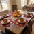 Fountain Leaf Placemat in Burgundy by Carole Shiber, a fall-themed dining table setting with autumn leaves shaped burgundy placemats, featuring dinnerware, glassware, and a centerpiece of pumpkins and dried leaves.