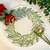 Add a Wreath-on-Round, 15" Placemat Carole Shiber Designs, decorative placemat featuring a painted green leafy wreath with red and yellow berry accents and holiday embellishments.