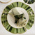 Add a Shapely LilyPad Placemat Carole Shiber Designs, a green leaf-shaped decorative placemat with gold trim under a white dinner plate and table setting.