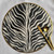 Zebra on the Round Carole Shiber Designs, a round decorative plate with a black and white zebra stripe pattern and a gold rim, accompanied by gold utensils.