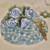 Classic Hydrangea Napkin Bouquet for 4, elegant table setting with white napkins shaped as roses with blue accents, green leaf details, placed on floral themed placemats, accompanied by golden cutlery and a wine glass.
