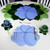 Buttercup Placemat - Light Blue Carole Shiber Designs, decorative table setting with blue flower-shaped placemat and matching napkin, wrapped in a blue flower-shaped napkin ring, silverware, and green floral accents on a white tablecloth.