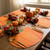 Painterly Pumpkin Placemat by Carole Shiber, autumn-themed dining table setting with pumpkin-shaped placemats, decorative pumpkins, faux leaves, candles, and elegant gold utensils for fall celebration.