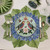 Carole Shiber Hydrangea Pacemat, a decorative table setting with a floral-themed plate featuring birds and butterflies, placed on a green and blue floral placemat with silverware, a glass, and a napkin.
