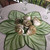 30" Tropical Linen Topper Carole Shiber Designs, decorative table centerpiece with large green and white tropical leaf design and cream-colored roses with green accents. 30" Tropical Linen Topper Carole Shiber Designs, decorative table centerpiece with large green and white tropical leaf design and cream-colored roses with green accents.