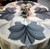 Silver & Grey Set of 10 Carole Shiber Designs, decorative tablecloth with a floral pattern in shades of silver, grey, and cream, topped with a matching floral centerpiece.