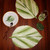 Little Minty Leaf Coasters, S/4 Carole Shiber Designs, decorative coasters featuring green leafy design on white background, set of four, displayed on a wooden table with plate, spoon, glass, and large leaves.