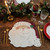 Santa Claus Placemat by Carole Shiber, festive shaped placemat featuring a cheerful Santa face with a white beard and red hat, styled on a Christmas table with greenery, candles, and gold cutlery.
