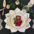 White/Silver Poinsettia Place-Setting Carole Shiber Designs, elegant holiday table setting with a poinsettia-themed napkin on a white plate with gold trim, surrounded by silverware, glassware, and festive greenery.