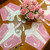 Vintage Party Gift Box - Pink/White Carole Shiber Designs, pink and white floral napkins with a bouquet of pink paper roses, and elegant glassware on a wooden table.