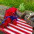 Stars & Stripes Napkin Bouquet by Carole Shiber Designs, a patriotic table decoration with red and blue fabric flowers on a red-and-white striped napkin, alongside a wooden planter with green plants and a basket holding silverware.
