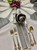 White/grey Wedding Centerpiece Carole Shiber Designs, elegant tabletop setting with floral centerpiece, folded napkin rose, gold and silver cutlery, and floral accents.