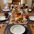 Thanksgiving Bouquet Carole Shiber Designs, an autumn-themed table setting with floral arrangements resembling roses in warm colors placed in wine glasses, surrounded by pumpkins, candles, and fall decor.
