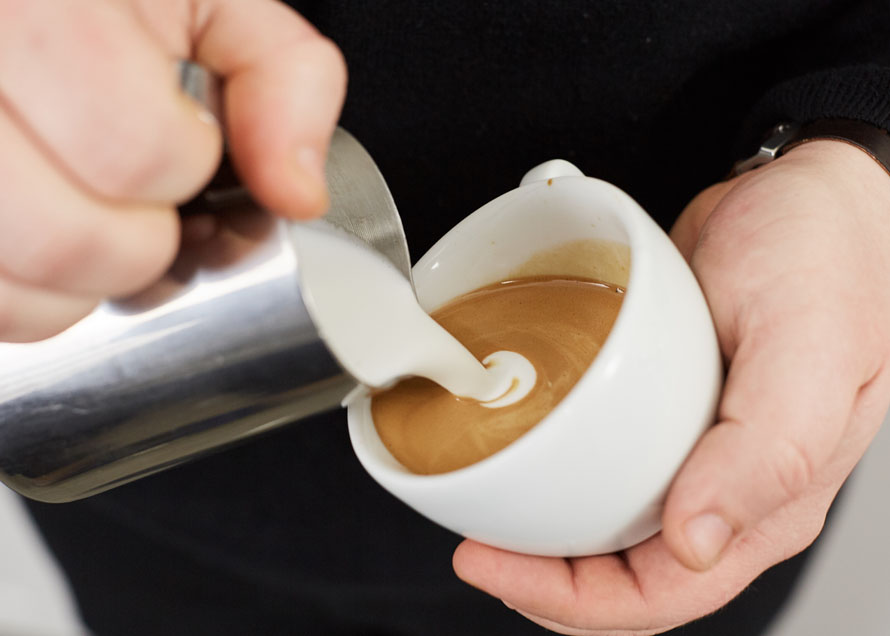 Pouring milk from a steaming pitcher at the surface of the beverage