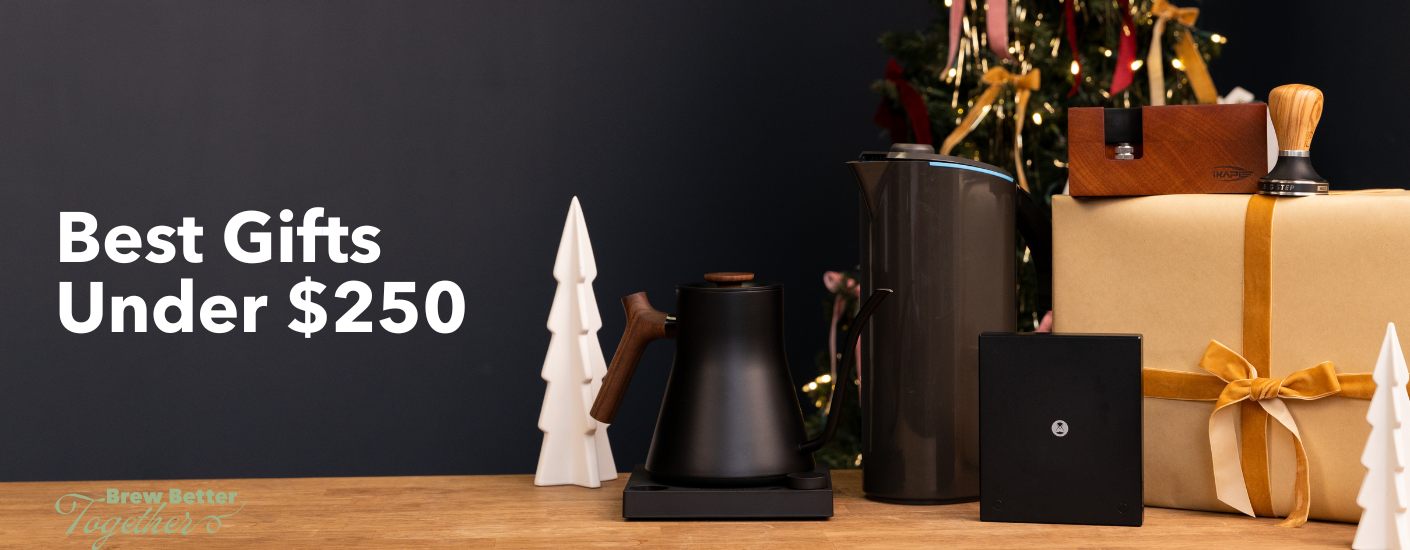 Fellow EKG Stagg kettle, Peak Water Pitcher, Timemoe Scale, Norcome knockbox and Pullman tamper sitting on a table with a wrapped gift and a Christmas themed background.